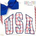 USA Plaid Filled Embroidery Design: The word USA in red gingham collegiate applique letters with a navy outline on a white shirt with a blue bow