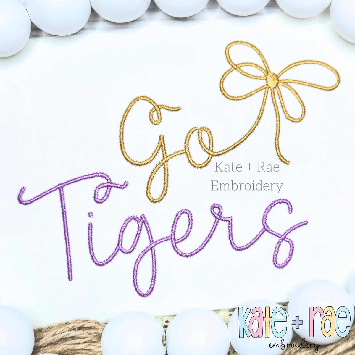 Preppy Go Tigers Embroidery Design: The words Go Tigers in gold and purple script with a gold bow on white fabric