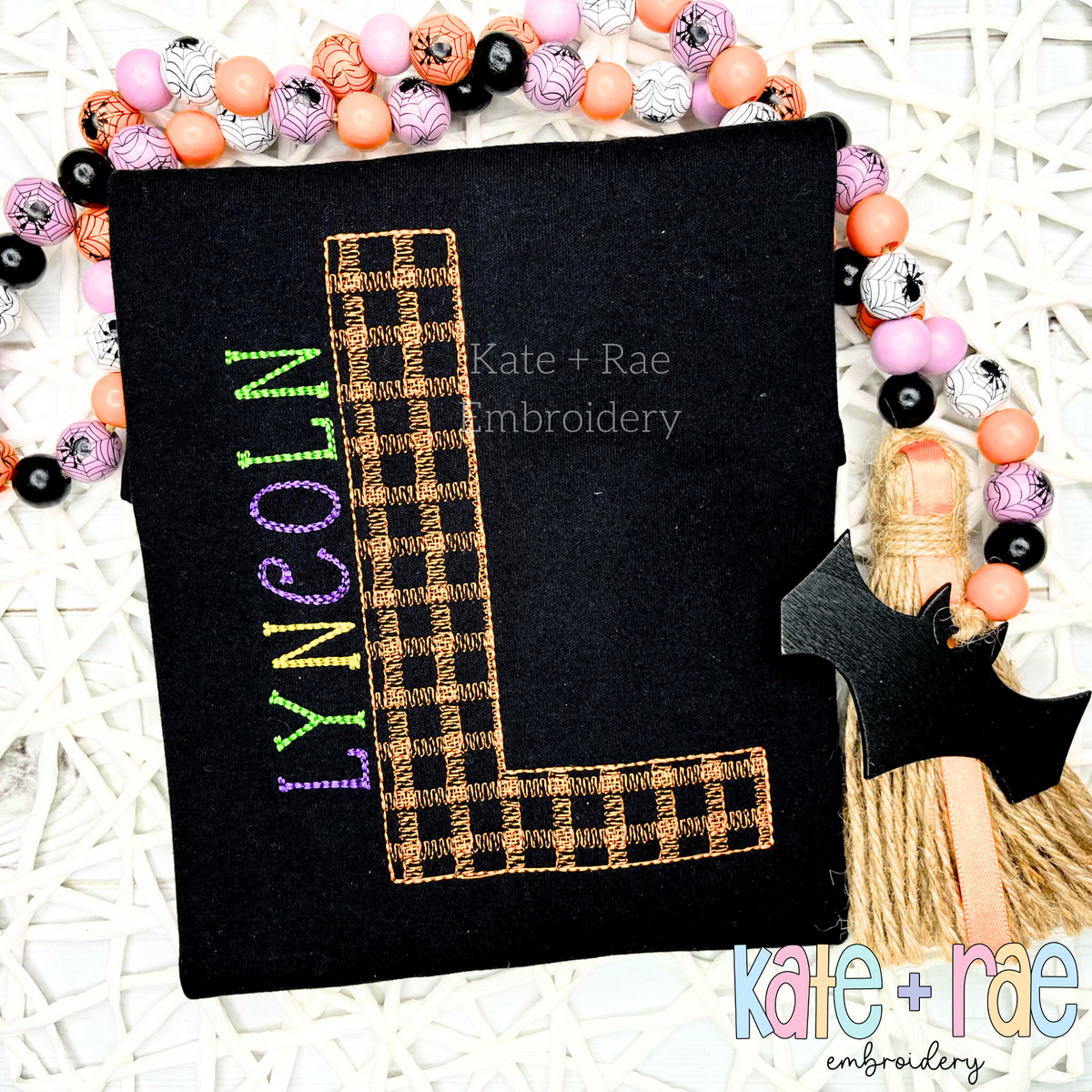 Plaid Embroidery Font: Orange and black plaid letter L with the name LYNCOLN in green and purple on a black shirt