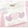 Heart Trio Motif Fill Stitch Embroidery Design: Three motif fill hearts in purple and pink above the name Sadie Rae on a white shirt over pink seersucker