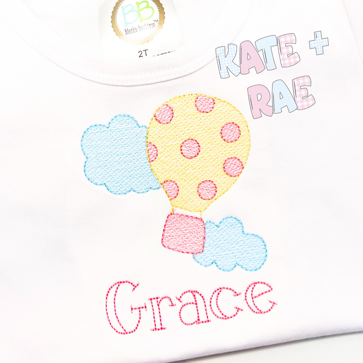 Hot Air Balloon Sketch Stitch Embroidery Design: Yellow polka dot hot air balloon between blue clouds above the name Grace in pink on a white shirt