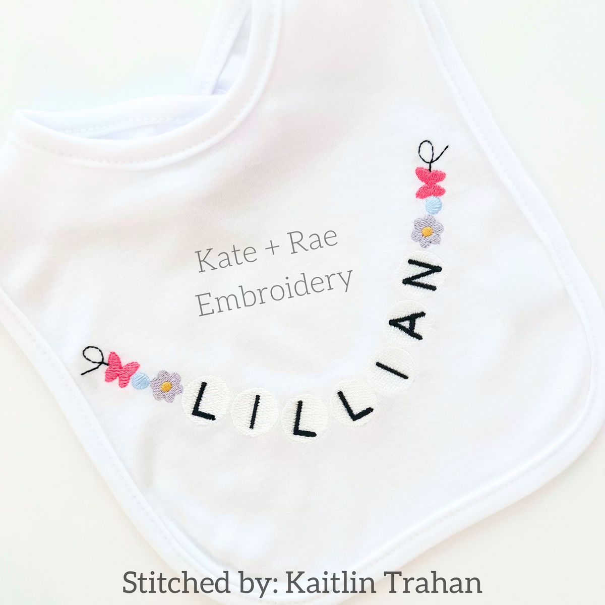 Friendship Embroidery Font: Friendship bracelet style beaded letters spelling Lillian around the neckline of a white baby bib