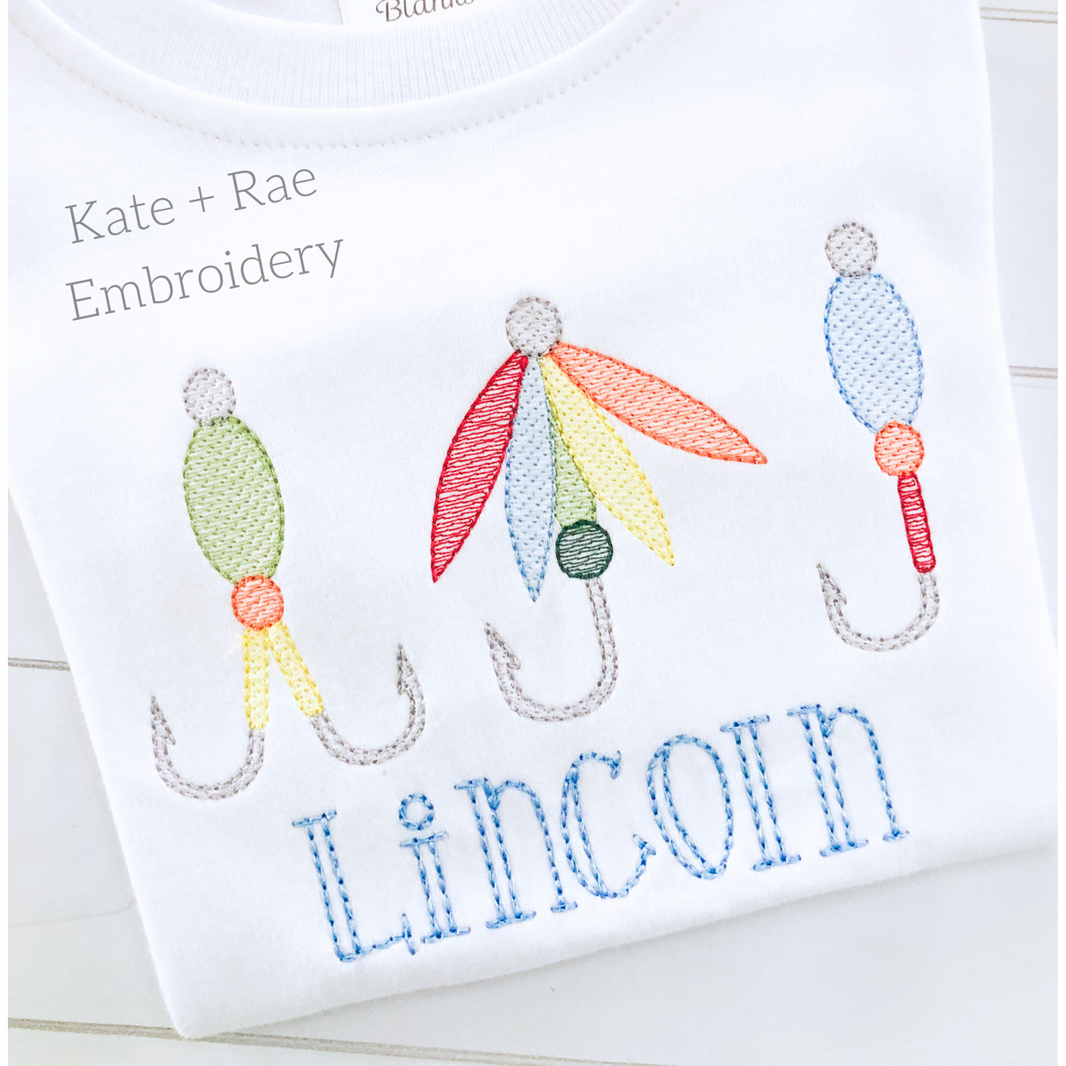 Fishing Lure Trio Sketch Stitch Embroidery Design: Three sketch stitch fishing lures above the name Lincoln in blue on a white shirt
