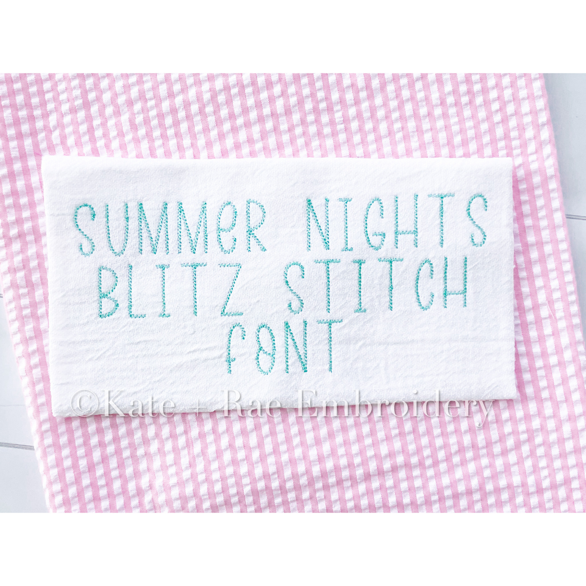 Summer Nights Blitz Stitch Embroidery Font: The words Summer Nights Blitz Stitch Font stitched in teal on white fabric over pink seersucker