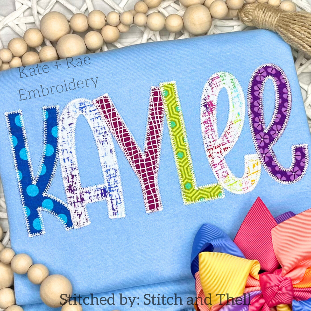 Kinley Zigzag Stitch Applique Embroidery Font: The name Kaylee in multicolored patterned applique letters on a light blue shirt with a bow