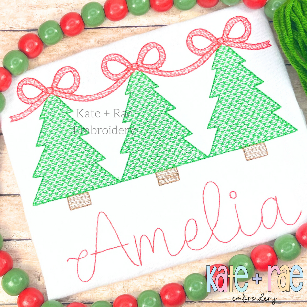 Girls Motif Fill Christmas Tree Trio with Connecting Bows Embroidery Design: Three green motif fill Christmas trees linked by red bows above the name Amelia in red script on a white shirt