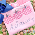 Girl's Plaid Filled Pumpkins with Bows Sketch Embroidery Design: Three pink plaid pumpkins with bows above the name delaney in gray script on a pink shirt