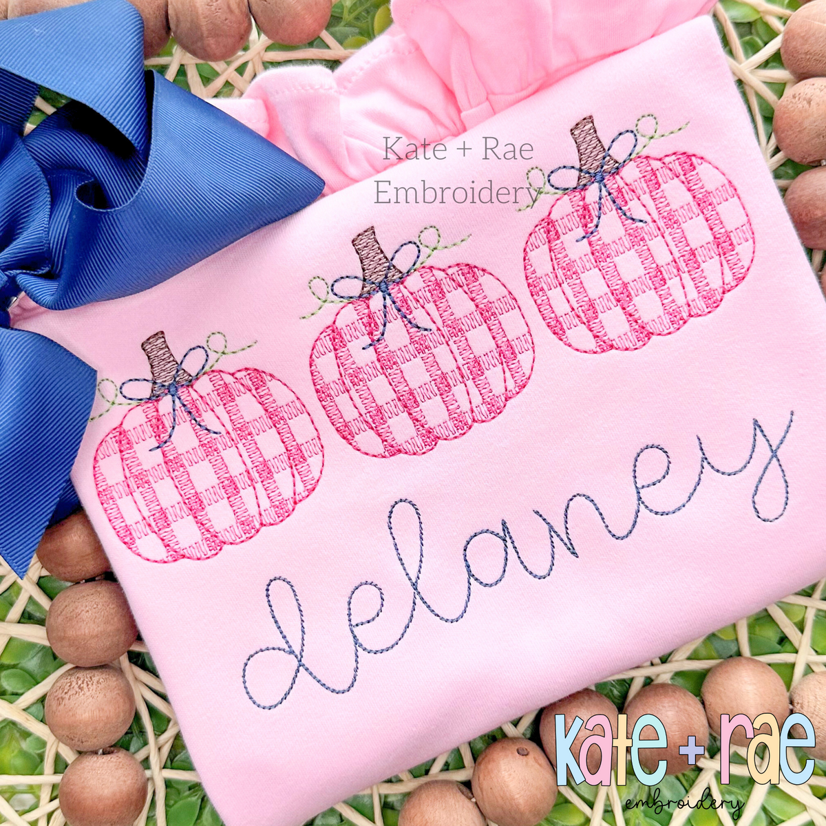 Girl's Plaid Filled Pumpkins with Bows Sketch Embroidery Design: Three pink plaid pumpkins with bows above the name delaney in gray script on a pink shirt