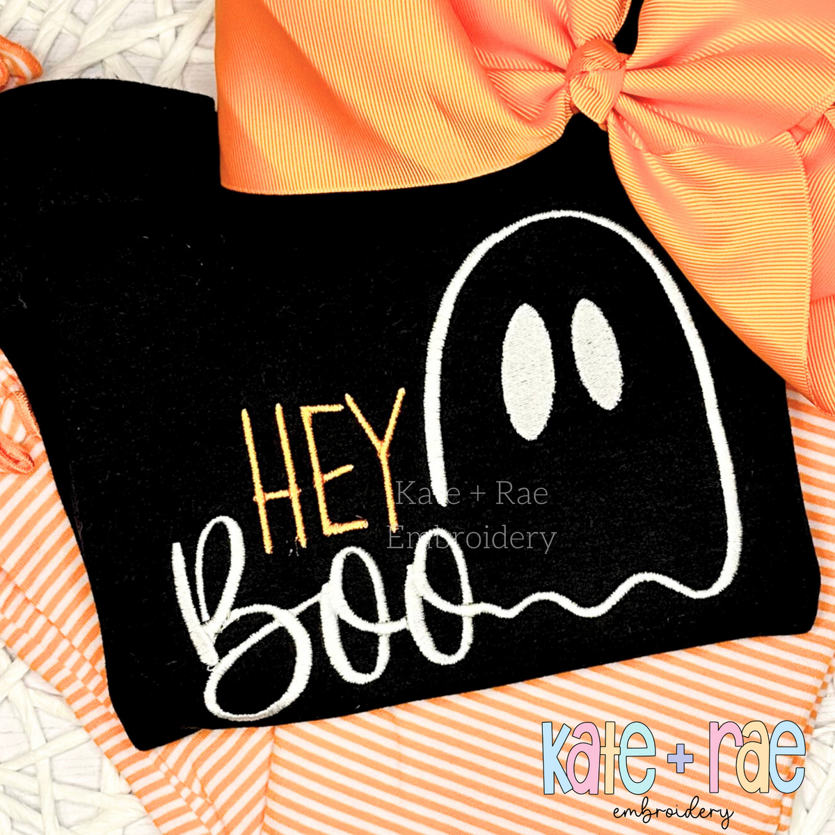 Hey Boo Halloween Embroidery Design: The words Hey Boo with a white ghost outline stitched on a black shirt with an orange bow