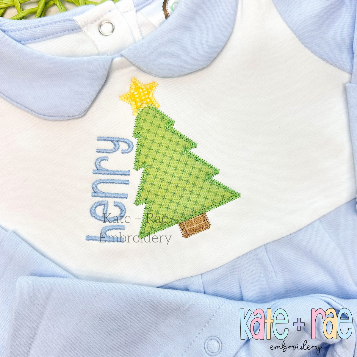 Christmas Tree with Stars Zigzag Applique Embroidery Design: Green Christmas tree applique with a yellow star beside the name henry on a white bib over a blue romper