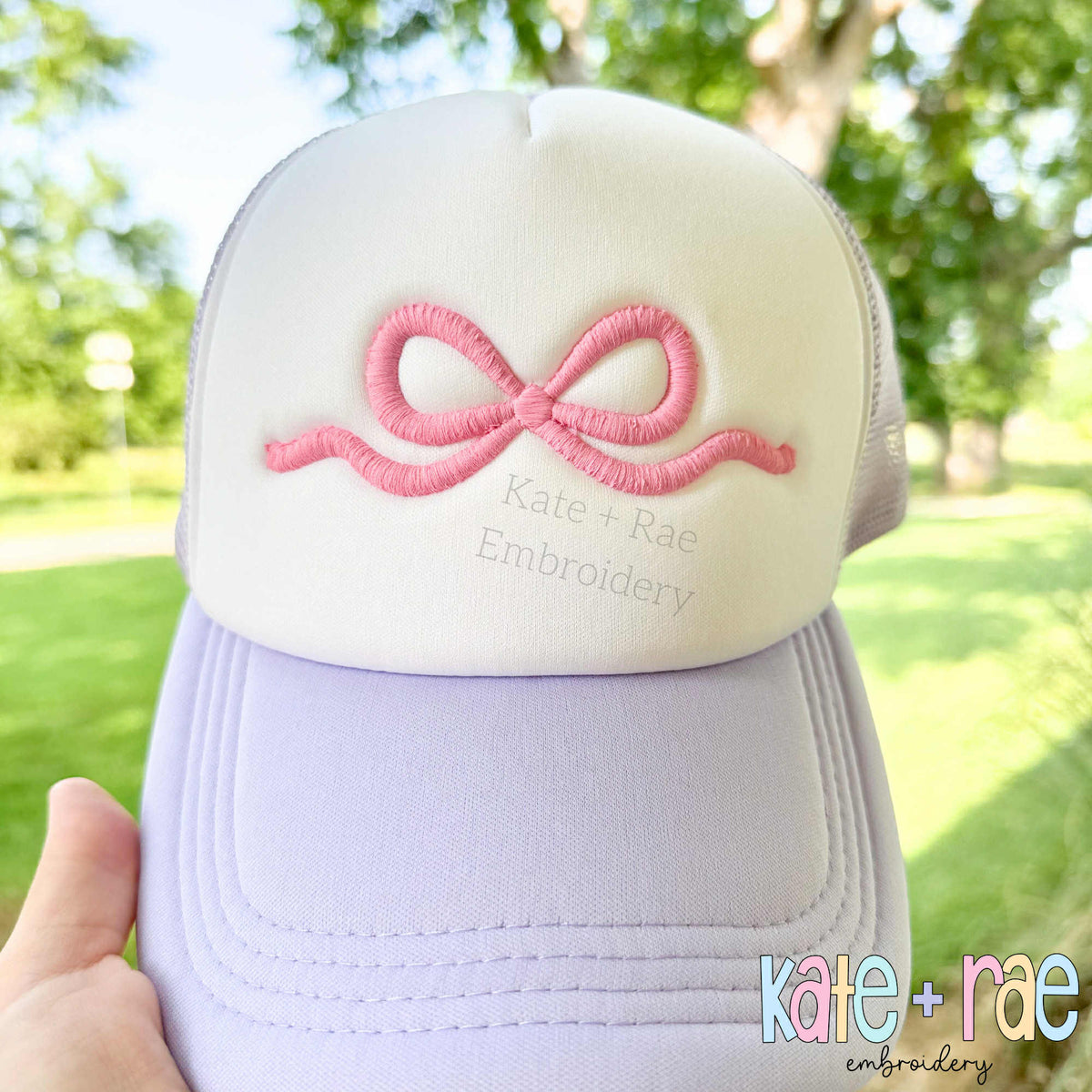 Flowy Bow 3D Puff Satin Stitch Embroidery Design: Pink 3D puff flowy bow on the front of a white and lavender trucker hat