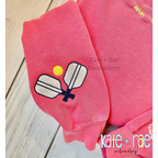 Pickleball Mini Fill Embroidery Design: Small crossed pickleball paddles with a yellow ball stitched on the sleeve of a coral sweatshirt