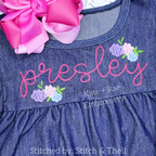 Kinley Floral Mini Fill Stitch Embroidery Design: The name presley in pink script with small pastel floral clusters on a chambray dress with a pink bow