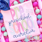 Preschool Sketch Stitch Embroidery Design: The words Pre School in pastel letters with preschool and the name aurelia in navy script on a pink shirt