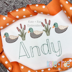 Mallard ducks and cattails in sketch stitch above the name Andy in green on a white shirt