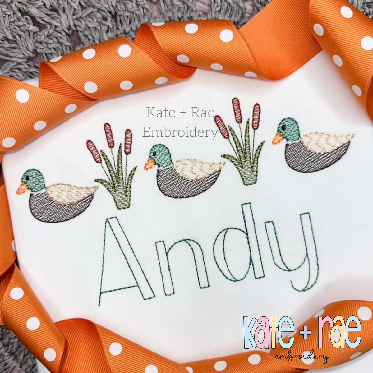 Mallard ducks and cattails in sketch stitch above the name Andy in green on a white shirt