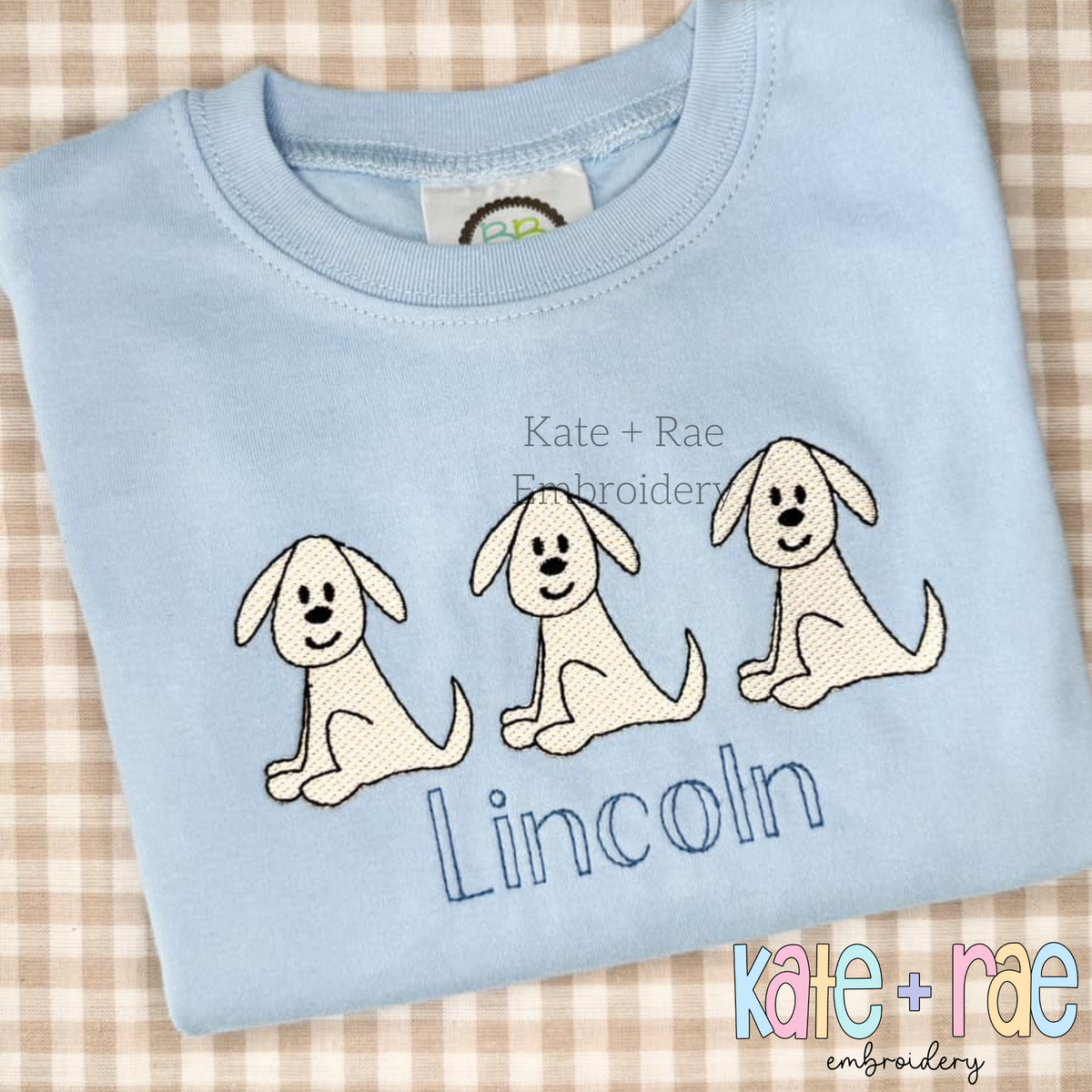 Three cream sketch stitch puppies above the name Lincoln in blue on a light blue shirt