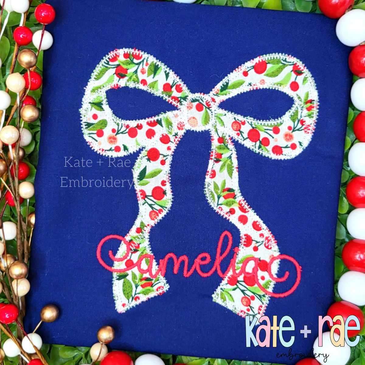 Merritt Bow Zigzag Stitch Applique Embroidery Design: Cherry print Merritt bow applique above the name Amelia in pink script on a navy shirt