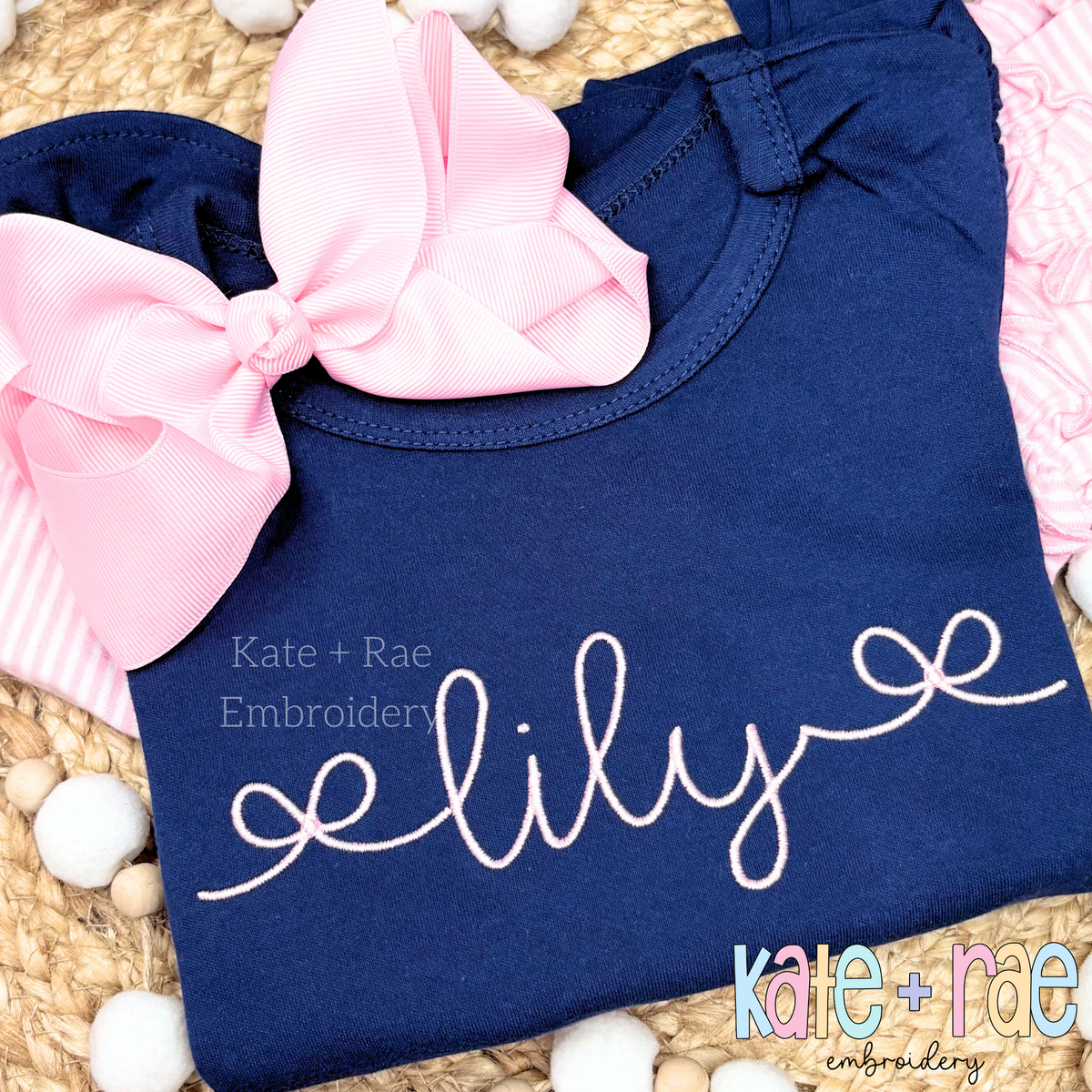 Bow Font Add On Embroidery Design: The name lily in pink script with curly bow ends on a navy shirt