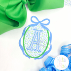 Oval Monogram Frame with Long Tail Bow Satin Applique Embroidery Design: Blue bow oval frame around a blue floral applique with a letter A on a white shirt