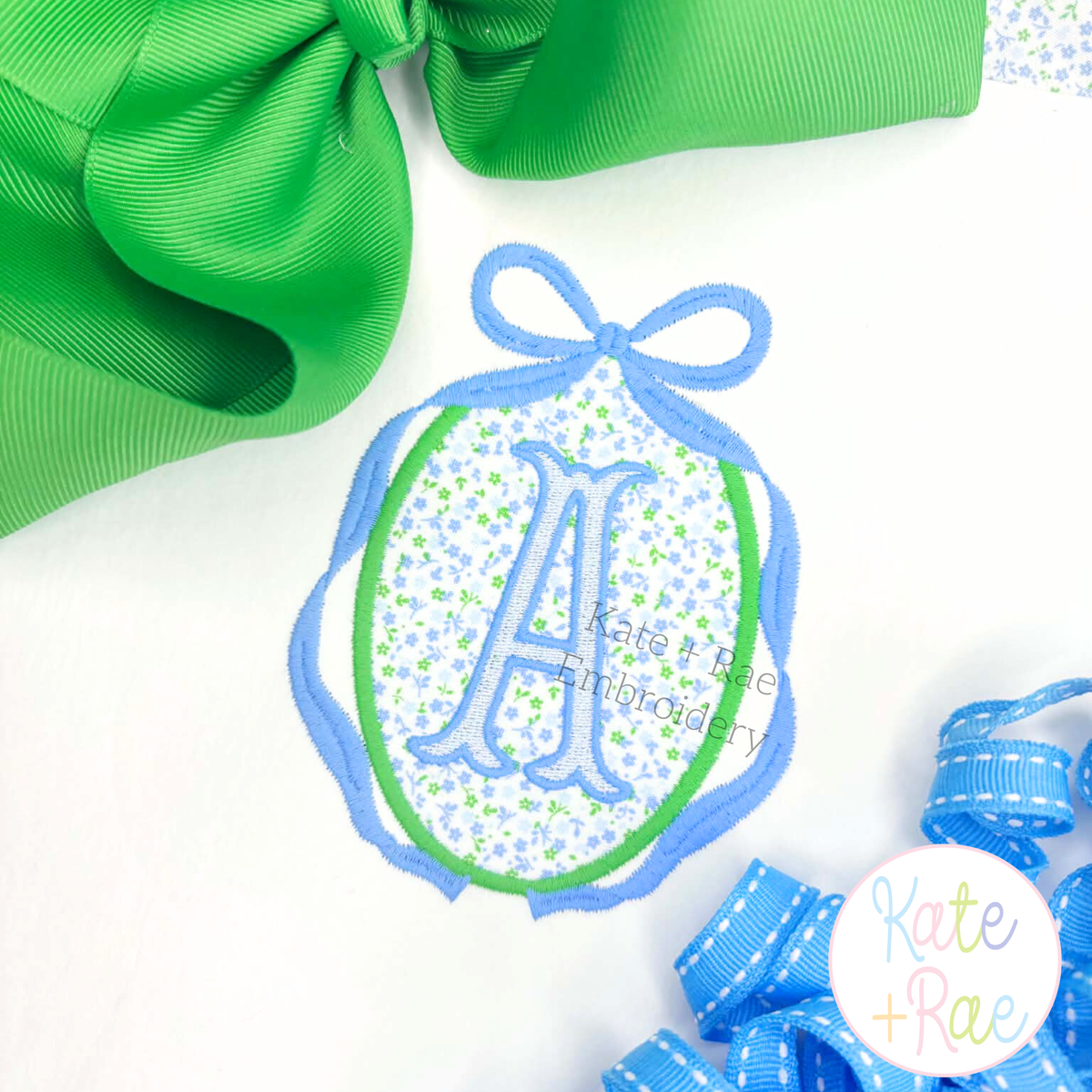 Oval Monogram Frame with Long Tail Bow Satin Applique Embroidery Design: Blue bow oval frame around a blue floral applique with a letter A on a white shirt