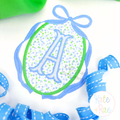 Oval Monogram Frame with Long Tail Bow Satin Applique Embroidery Design: Blue oval frame with long tail bow around a blue floral oval with a letter A on a white shirt