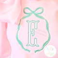 Emma Satin Stitch Long Tail Monogram Bow Embroidery Design: Mint long tail bow frame around a mint fishtail letter E on a pink shirt