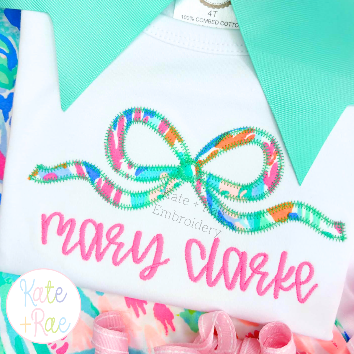 Flowy Longtail Bow Zigzag Stitch Applique Embroidery Design: Bright print flowy ribbon bow applique above the name mary clarke in pink script on a white shirt