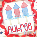 Girl's Firework Trio with Bows Sketch Stitch Embroidery Design: Three red and blue sketch firework rockets with light blue bows above the name Aubree in red on a white shirt