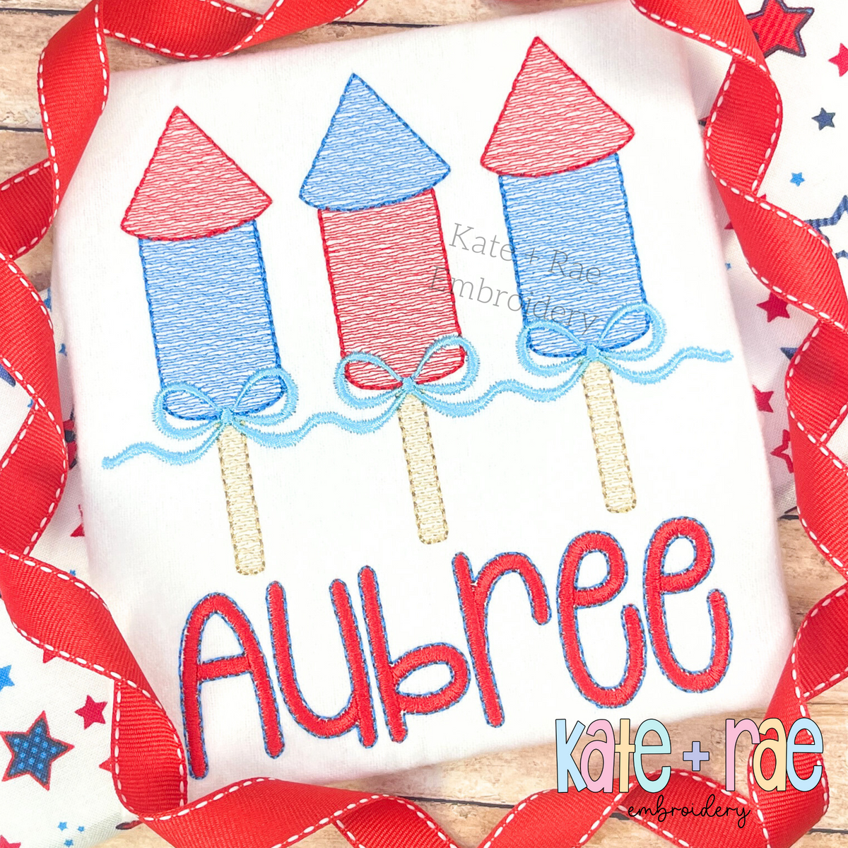 Girl's Firework Trio with Bows Sketch Stitch Embroidery Design: Three red and blue sketch firework rockets with light blue bows above the name Aubree in red on a white shirt