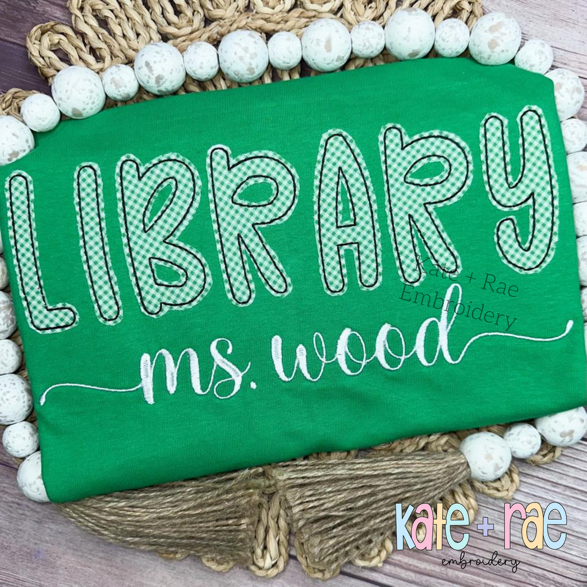 Playground Bean Stitch Applique Embroidery Font: LIBRARY in green patterned applique letters above ms. wood in white script on a green shirt