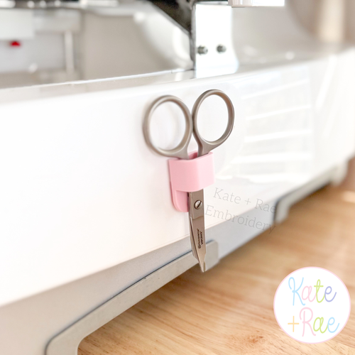 Adhesive Scissor Holders: Pink adhesive scissor holder holding scissors on the side of an embroidery machine