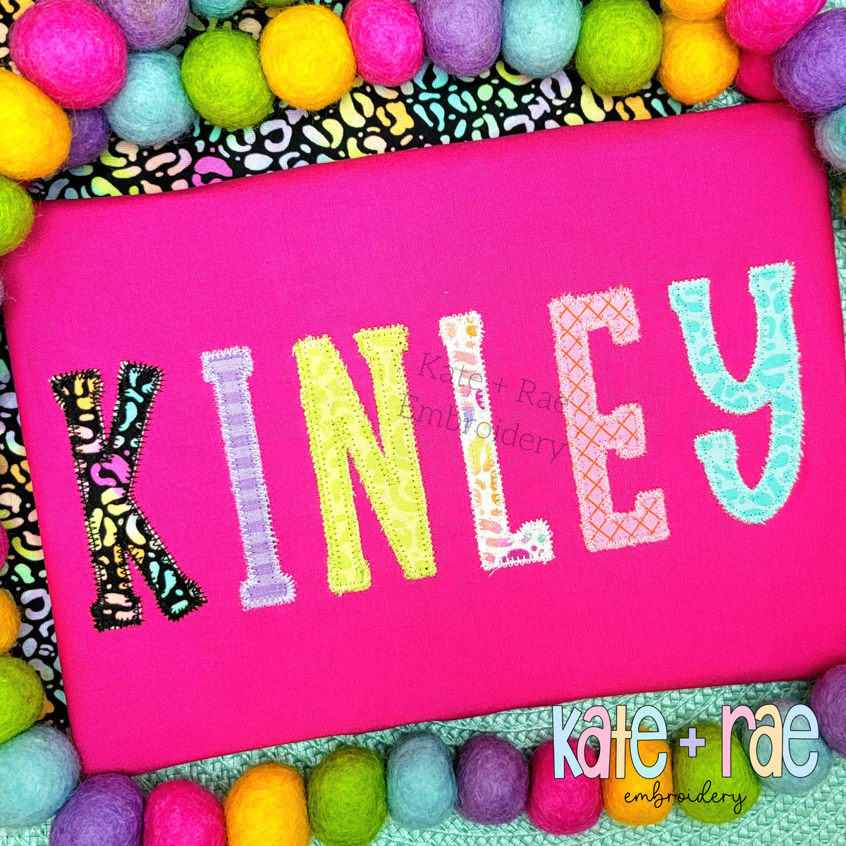 Gracie Zigzag Stitch Applique Embroidery Font: The name KINLEY in multicolor patterned zigzag applique letters on a hot pink shirt