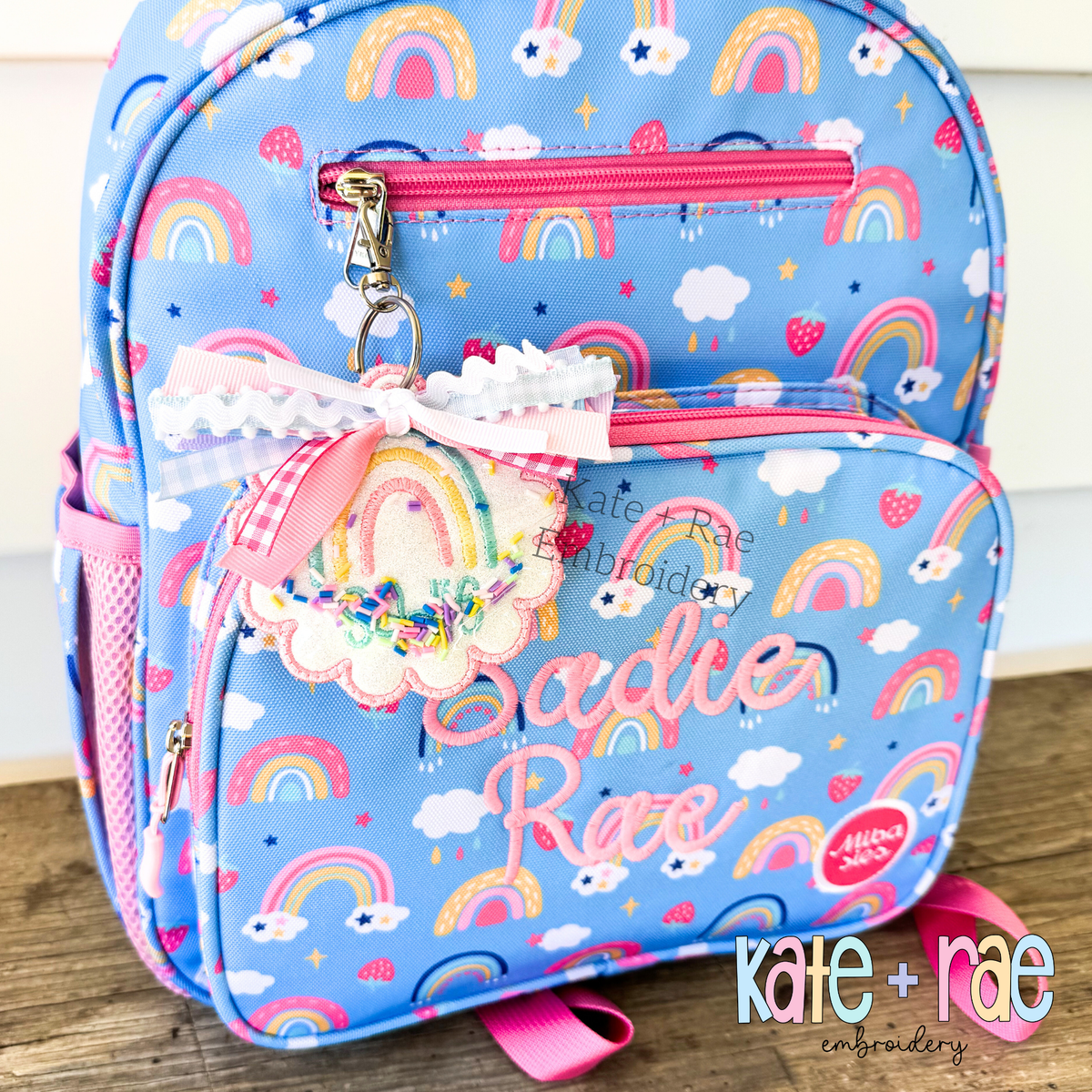 ITH Scalloped Circle Bag Tag Satin Stitch Applique Embroidery Design: Pink scalloped circle bag tag with a rainbow and confetti shaker window clipped to a rainbow backpack