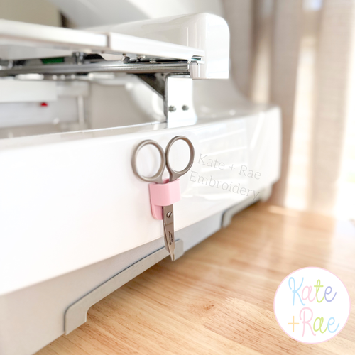 Adhesive Scissor Holders: Pink scissor holder mounted on a white embroidery machine with silver scissors