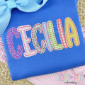 Playground Bean Stitch Applique Embroidery Font: The name CECILIA in patterned bean stitch applique letters on a royal blue shirt