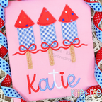 Girl's Firework Trio Zigzag Applique Embroidery Design: Blue gingham firework appliques with red tops and bows above the name Katie in red and blue on a pink shirt