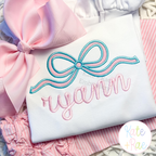 Flowy Longtail Bow Satin Stitch Applique Embroidery Design: Aqua and pink flowy bow satin applique above the name ryann in pink script on a white shirt