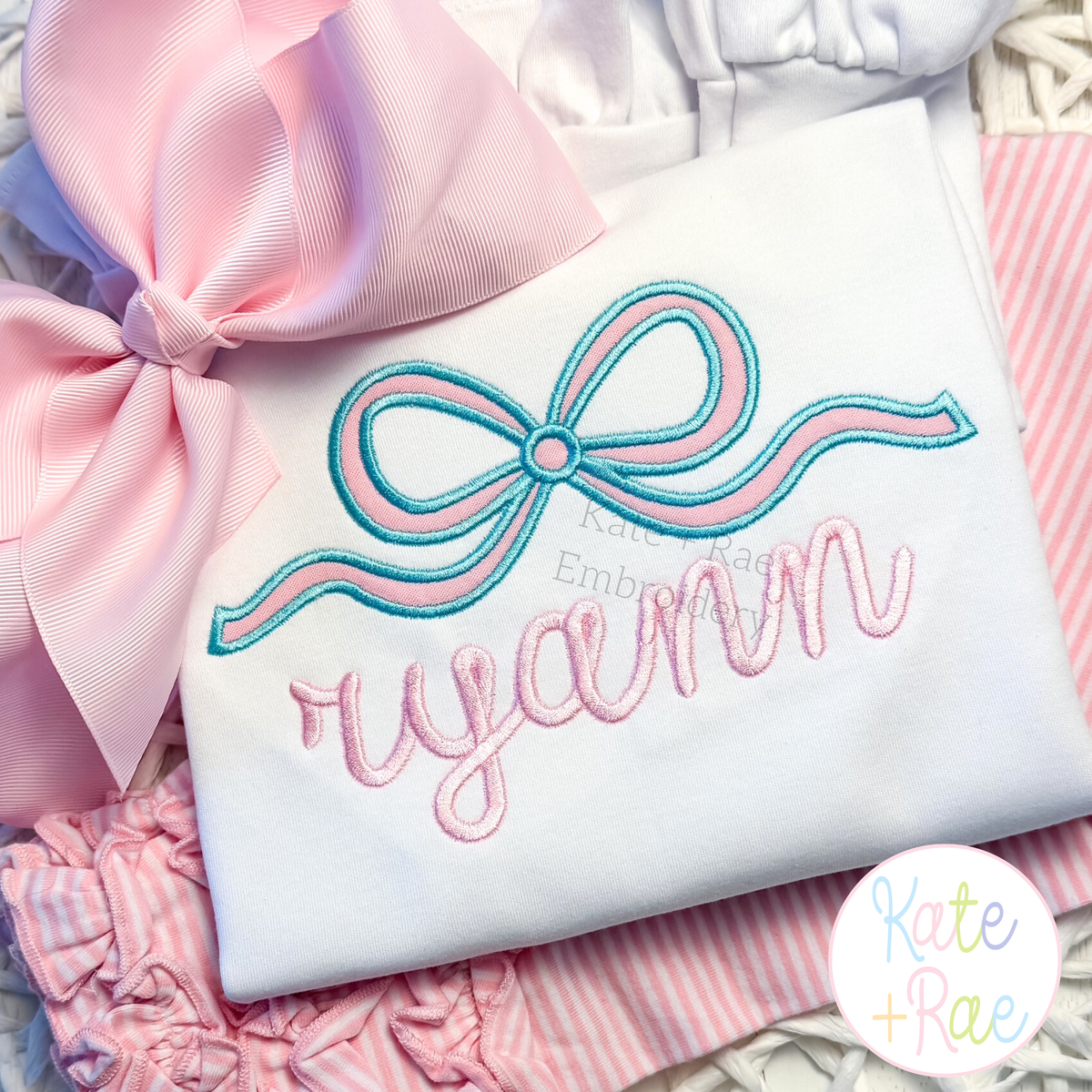 Flowy Longtail Bow Satin Stitch Applique Embroidery Design: Aqua and pink flowy bow satin applique above the name ryann in pink script on a white shirt