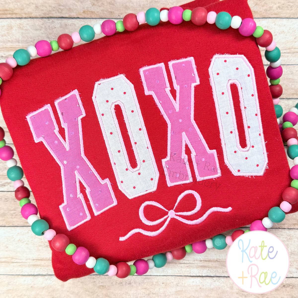 Curved XOXO with Bow Satin Stitch Applique Embroidery Design: The word XOXO in pink polka dot curved collegiate applique letters with a bow on a red sweatshirt
