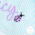 Ladybug Mini Fill Stitch Embroidery Design: Close-up of a purple mini ladybug beside a lavender script letter y on aqua seersucker
