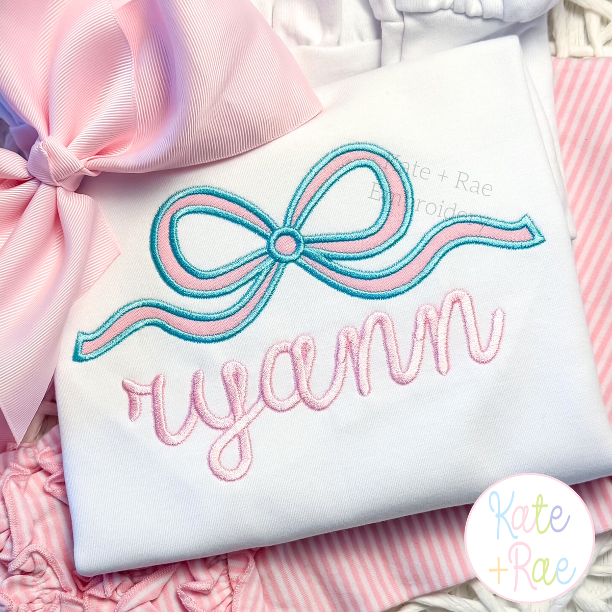 Flowy Longtail Bow Satin Stitch Applique Embroidery Design: Close-up of an aqua and pink flowy bow above the name ryann in pink script