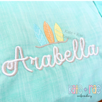 Delilah Two Toned Satin Stitch Embroidery Font: The name Arabella in two toned white satin script below three pastel surfboards on an aqua shirt