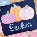 Girl's Fall Pumpkin Trio with Bows Zigzag Stitch Applique Machine Embroidery Design: Pink gingham, orange, and pink striped pumpkin appliques with blue bows above the name Decker on a navy shirt
