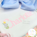 Floral Block Sketch Stitch Embroidery Font: The name Charlotte in pink sketch block letters with a tiny flower bouquet on a white shirt