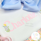 Floral Block Sketch Stitch Embroidery Font: The name Charlotte in pink sketch block letters with a tiny flower bouquet on a white shirt