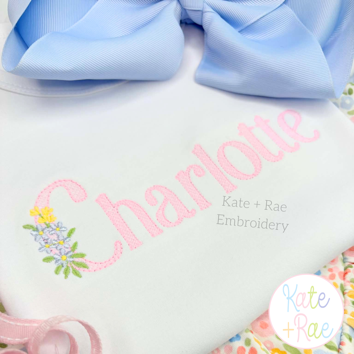 Floral Block Sketch Stitch Embroidery Font: The name Charlotte in pink sketch block letters with a tiny flower bouquet on a white shirt