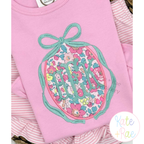 Oval Monogram Frame with Long Tail Bow Satin Applique Embroidery Design: Teal long tail bow oval frame around a pink floral applique monogram on a pink shirt