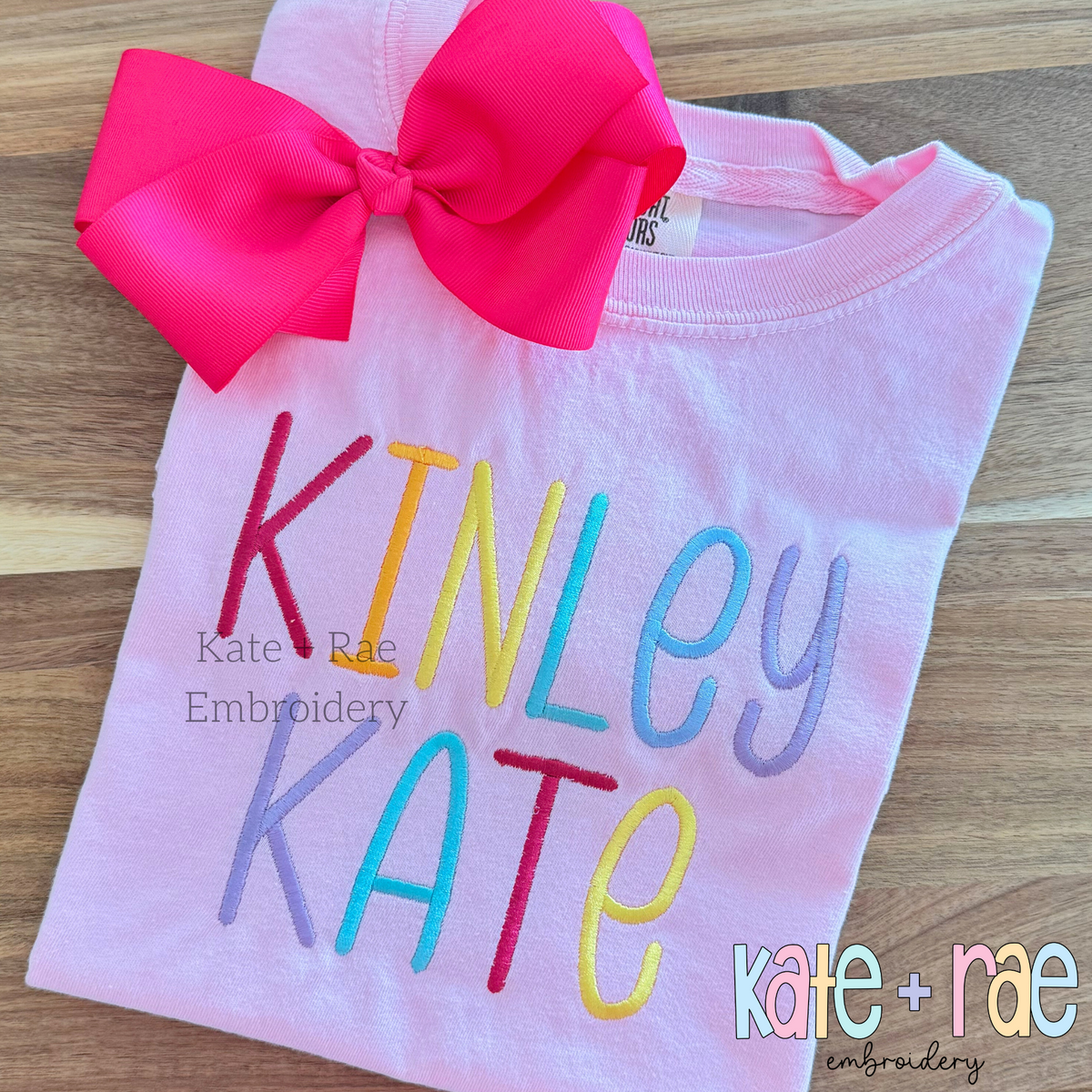 Summer Nights Satin Stitch Embroidery Font: The name Kinley Kate stitched in rainbow colored letters on a pink shirt with a hot pink bow