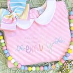 Flowy Bow Font Add On Embroidery Design: The name emily in rainbow script with flowy bow ends on a pink striped dress